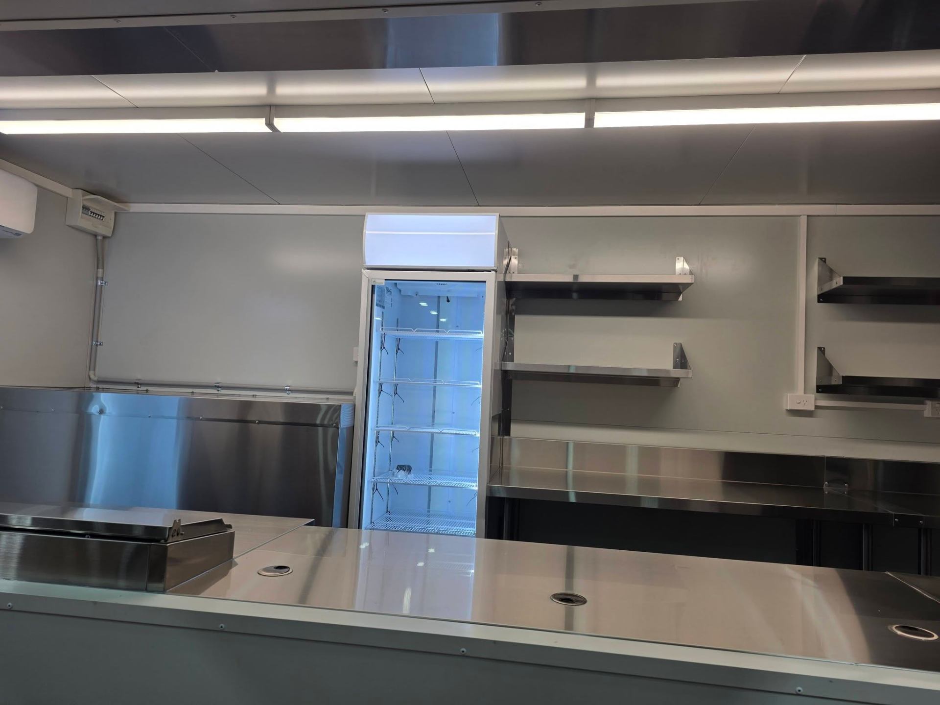 Interior fit out inside a Custom Food Vans build showing upright glass fridge, stainless wall shelving and serving counter